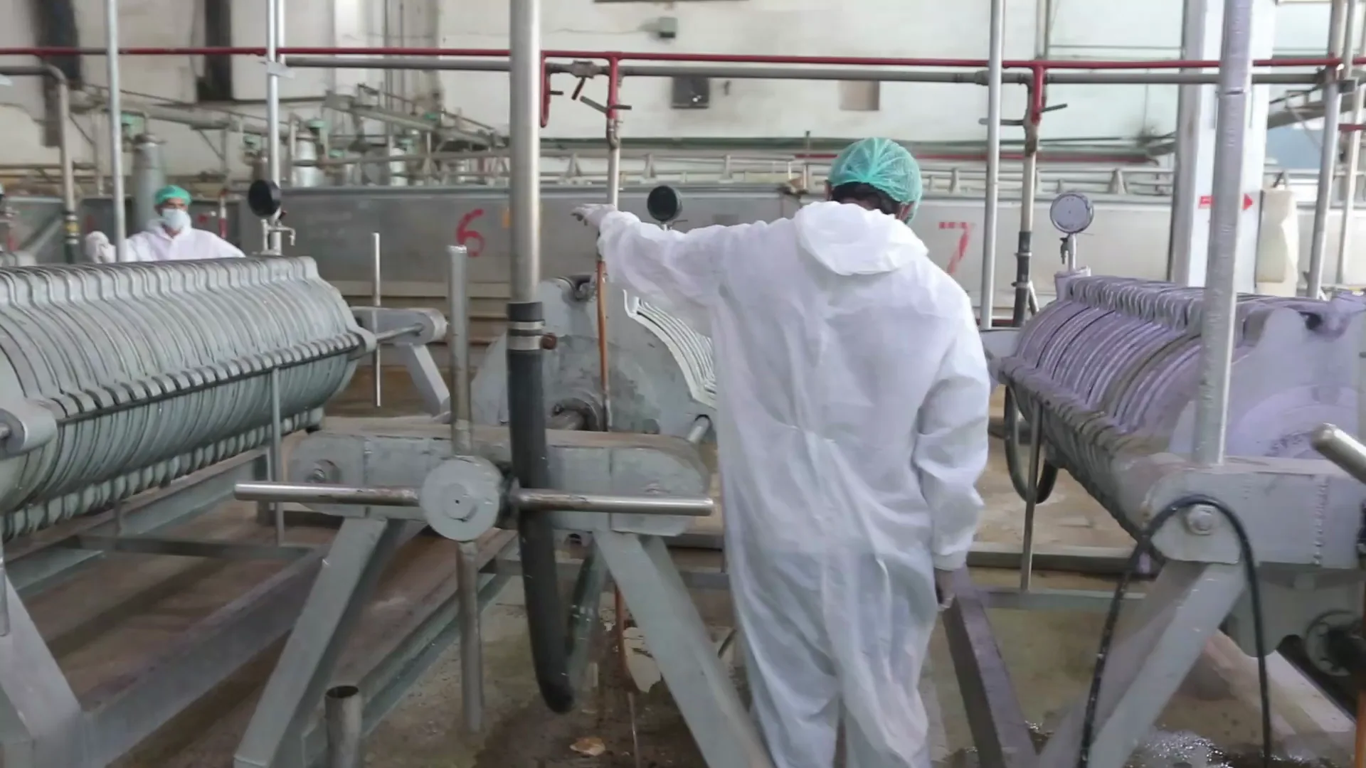 An operator in a white cleanroom suit walking between filter presses on the Sadiq Gelatin production floor.